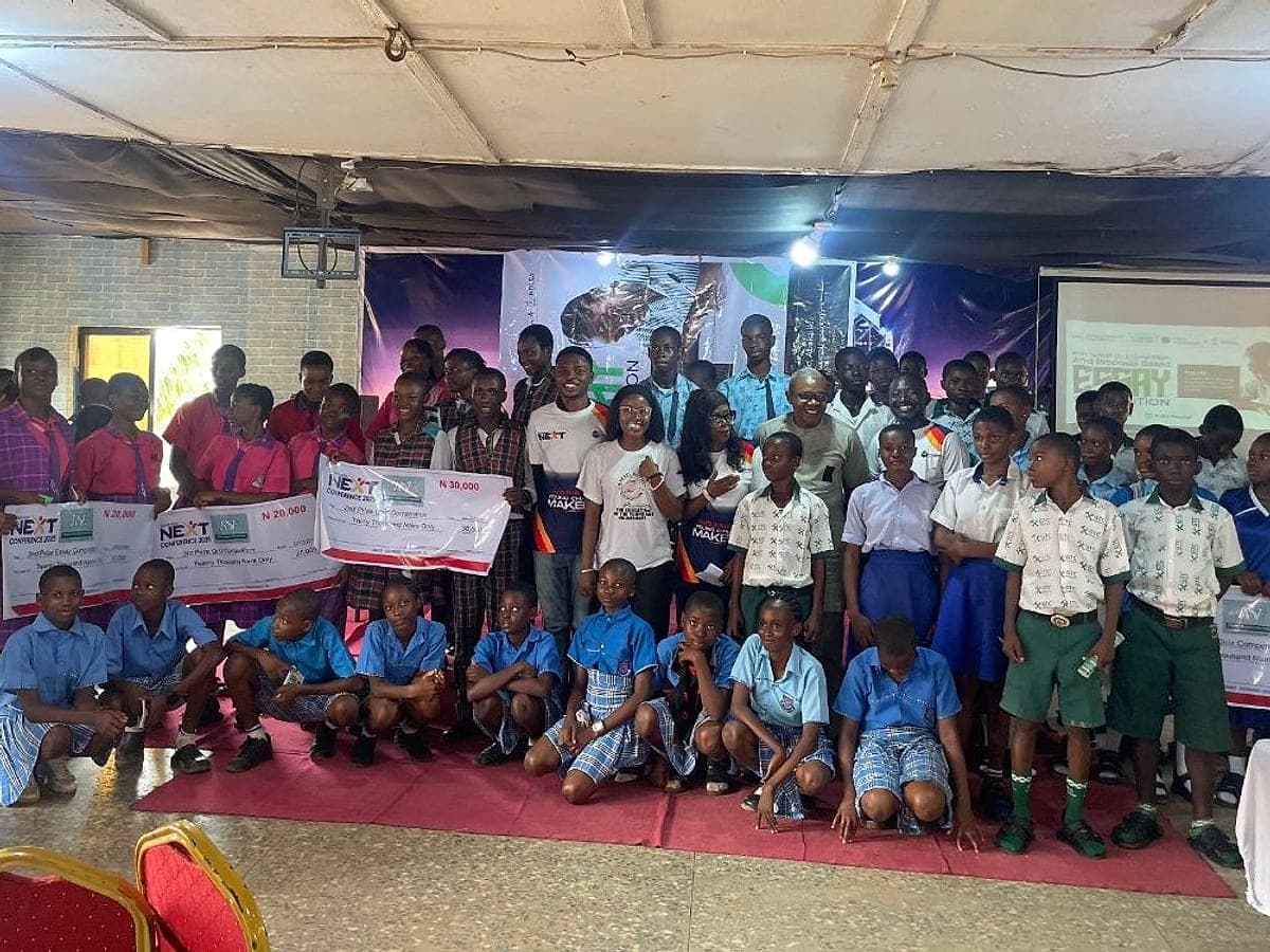 Pupils in school uniforms and RSF team members stand and kneel together on a red carpet at an education event, some holding large prize cheques, in front of a projector screen.