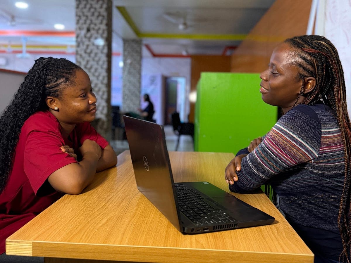 Two young women sit facing each other across a wooden table with a laptop between them, smiling as they talk one to one.