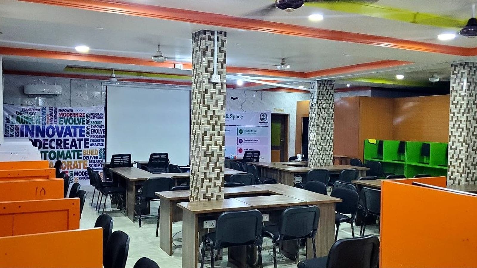 An empty, well-lit workspace with orange and green desk booths, wooden tables and a projector screen under a colourful ceiling.