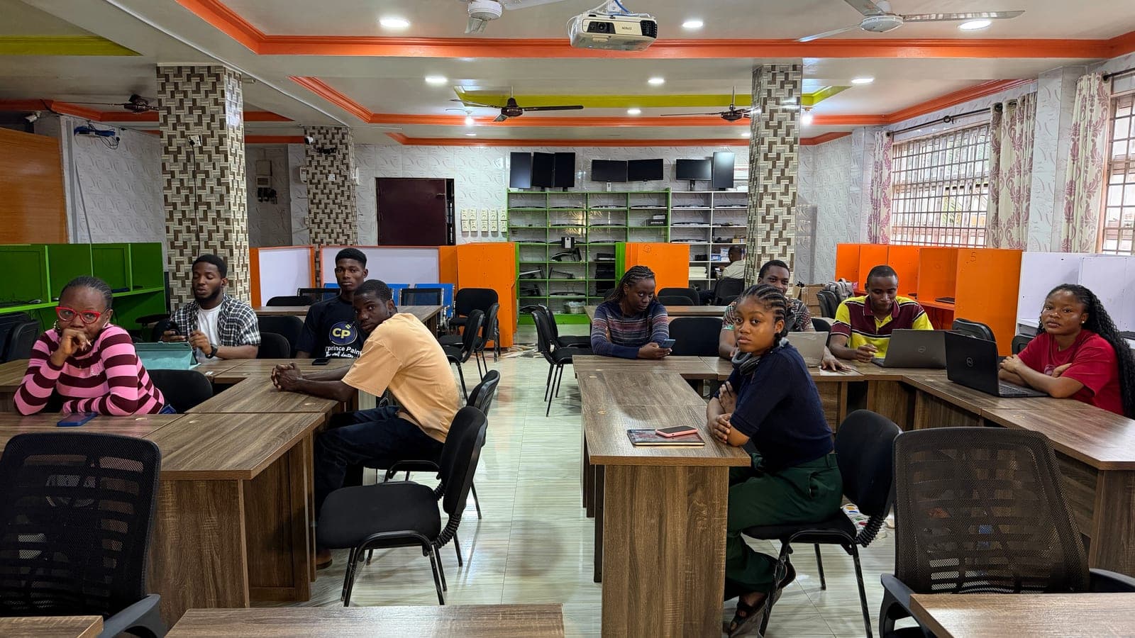 Learners sit at wooden desks in the training room, some with laptops and phones, looking towards the camera.