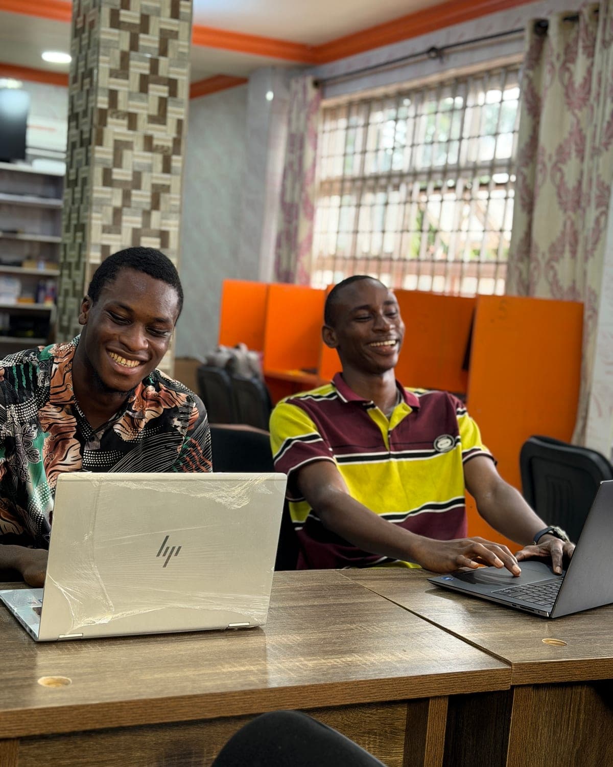 Two young men smiling as they work side by side on laptops at a desk in a bright co-working room.
